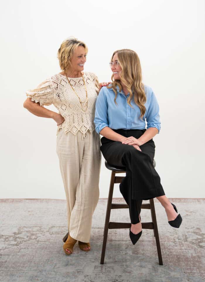 Two women smiling; one standing with hands on hips in a crochet top and beige pants, the other seated on a stool wearing glasses, a blue blouse, and black pants.