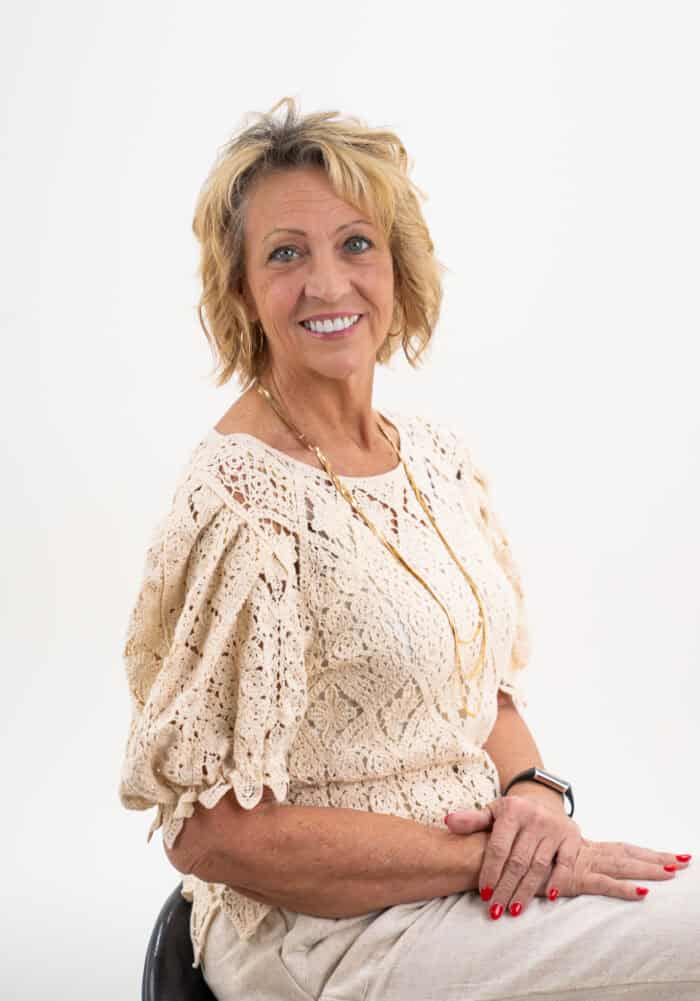 A woman with short blonde hair smiles while seated against a white background. She is wearing a beige lace top and a gold necklace, reflecting the warm charm of east Tennessee. Her hands rest on her lap, exuding grace and tranquility reminiscent of a serene countryside property.