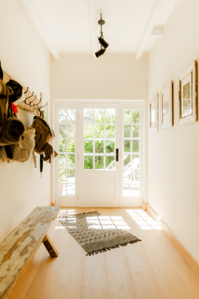 A bright entryway in this charming East Tennessee home features a glass door with sidelights, a wooden bench, a patterned rug, coat hooks adorned with jackets and bags, and framed pictures on beige walls. Sunlight filters through the door, creating an inviting space perfect for any homeowner.