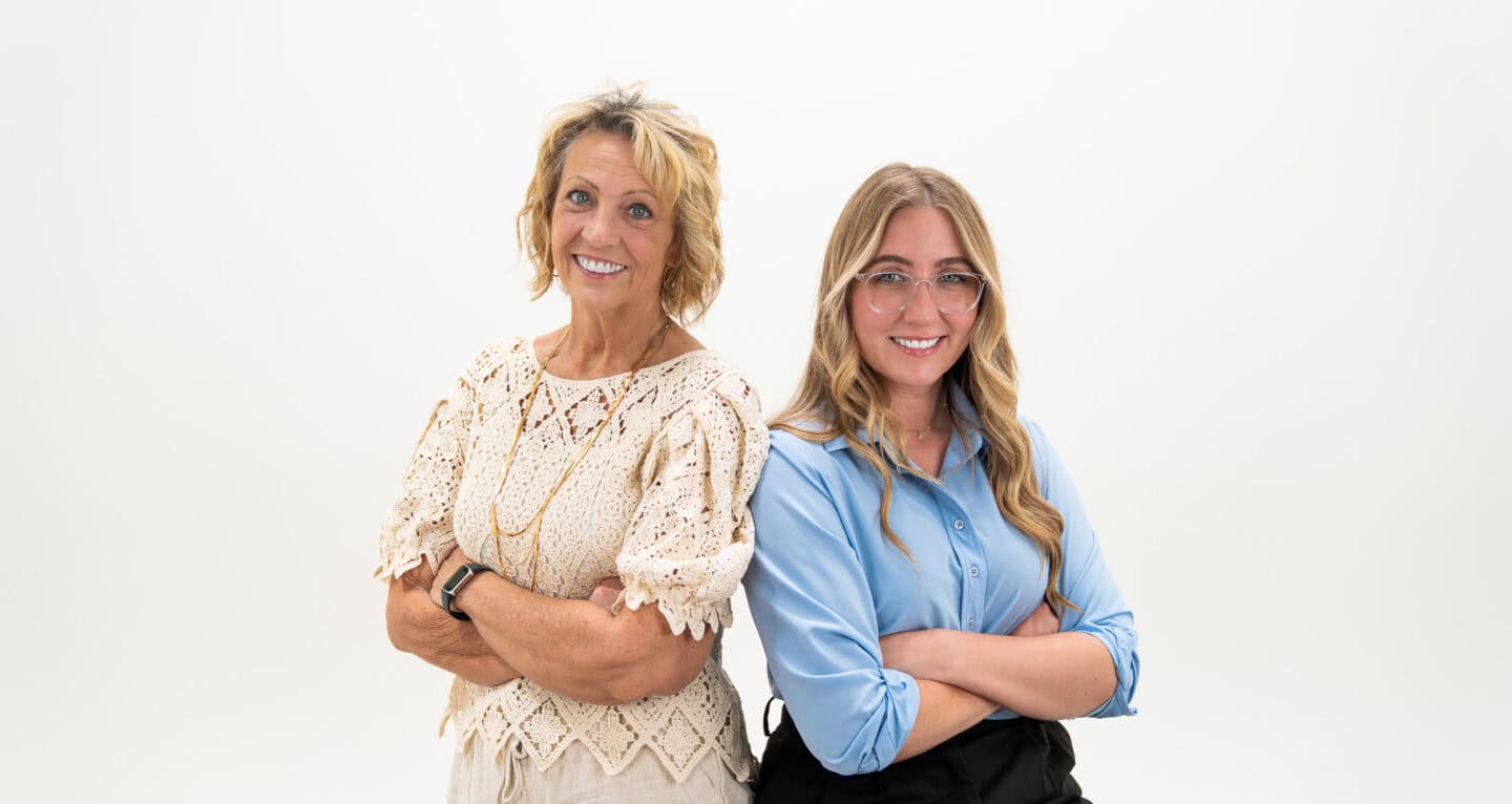 Two women stand back-to-back, beaming with confidence and arms crossed. The woman on the left wears a beige lace shirt, while the woman on the right sports a blue blouse. Perfect partners for any east Tennessee property management or real estate venture. White background.
